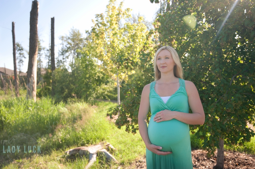 outdoor-maternity-portrait-wooded-area-one-hand-ontop-of-the-belly-and-one-hand-below-eyes-glancing-off-into-the-distance-sun-flare-in-the-right-corner-of-the-image