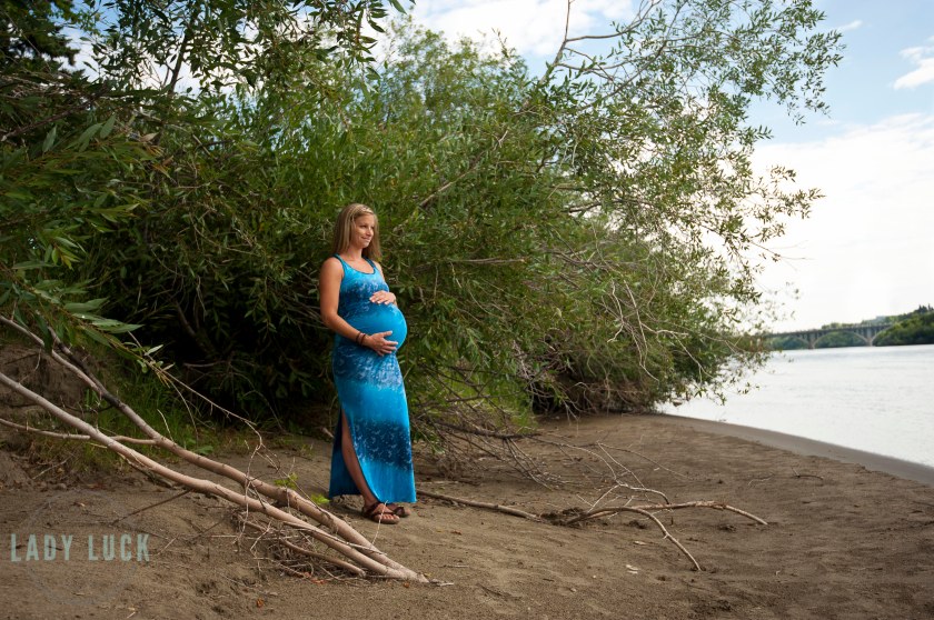 outdoor,maternity,session,in,saskatoon,along,the,river,bank,outside,the,delta,bessborough,client,wearing,a,blue,sarong,and,sandals,holding,her,belly,and,looking,off,toward,the,river,large,trees,in,the,background